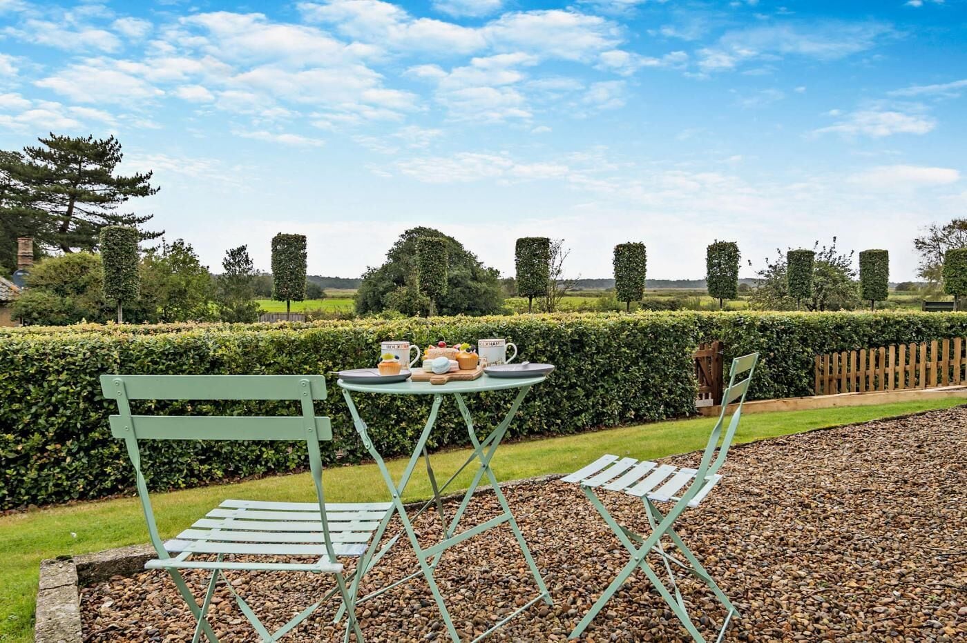 A garden setting with a table and chairs, a hedge, and a wooden fence.