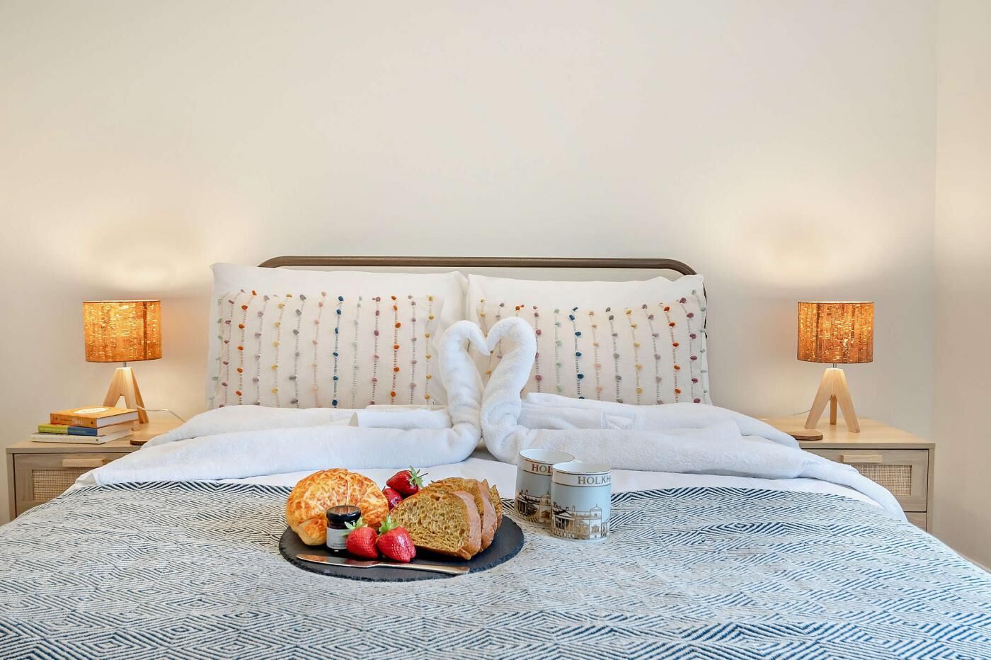Een netjes opgemaakt bed met een witte deken, twee kussens en een hartvormig kussen. Twee nachtkastjes met lampen en een dienblad met brood, aardbeien en jam.