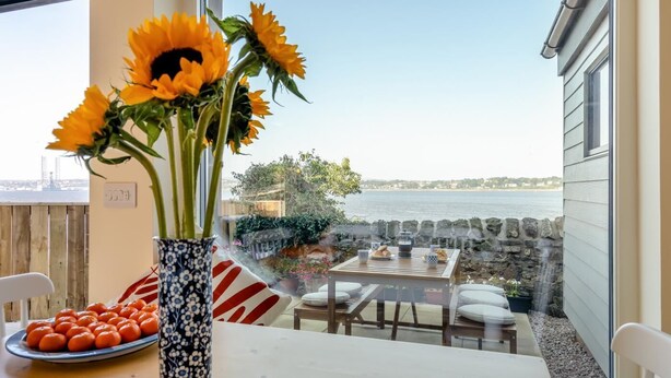 A dining area with a table set for two, a vase with sunflowers, and a view of the sea. - 2 Bed in Tayport (oc-w30005) (Dundee)