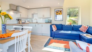 A modern kitchen with white cabinets, a dining area with a wooden table and white chairs, and a blue sofa with colorful cushions. - 2 Bed in Tayport (oc-w30005) (Dundee)