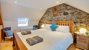 A bedroom with a stone wall, a wooden bed, a window, a bedside table, and a chair.