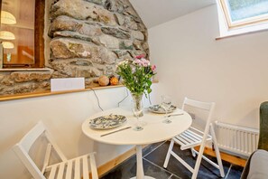A dining area with a stone wall, white chairs, a round table with plates and glasses, and a vase with flowers.