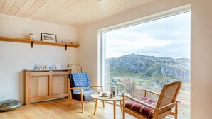 A cozy room with a wooden ceiling, a large window overlooking a mountainous landscape, a wooden cabinet with artwork, and a seating area with a table and chairs. - 2 Bed in Lochinver (oc-t30029) (Clachtoll)