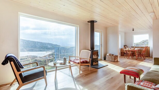 A spacious living room with a large window offering a view of a coastal landscape, a built-in wood-burning stove, and a dining area with wooden furniture. - 2 Bed in Lochinver (oc-t30029) (Clachtoll)