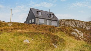 A modern house with a sloped roof, large windows, and a stone wall in the background. - 2 Bed in Lochinver (oc-t30029) (Clachtoll)