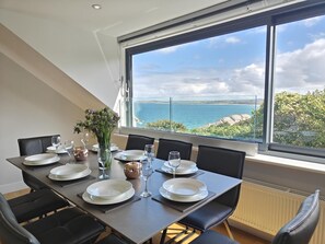 A dining area with a table set for six, overlooking a scenic view of the sea and greenery. - 4 Bed in Carbis Bay (oc-m30075) (Carbis Bay)