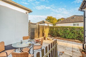 Een terras met een glazen tafel, rieten stoelen, een haag en een wit gebouw.