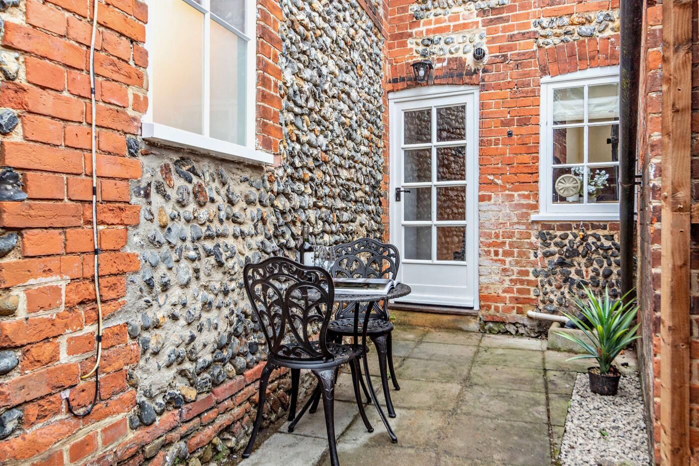 A brick building with a stone wall, a white door, and a window. There is a table and two chairs outside.