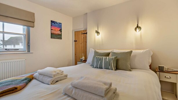A neatly made bed with folded towels, two wall-mounted light fixtures, a window with a view, and a door with a wooden frame. - 2 Bed in Lavenham (oc-s30055) (Lavenham)