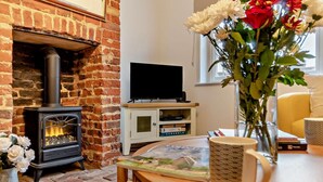 A cozy living room with a brick fireplace, a television on a wooden stand, a table with a mug, and a vase with flowers. - 2 Bed in Lavenham (oc-s30055) (Lavenham)