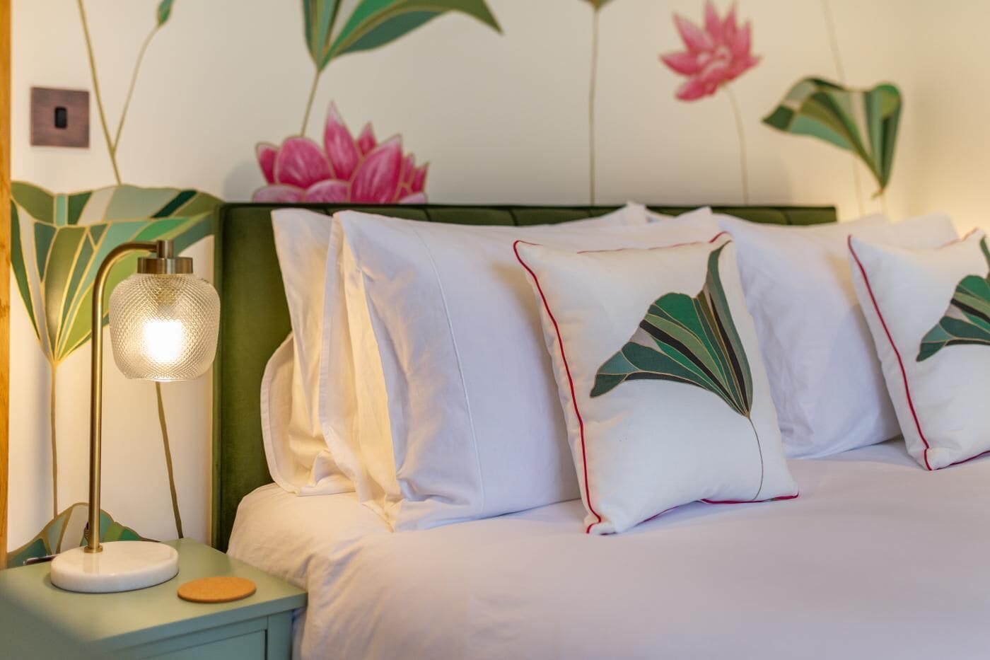 Ein ordentlich bezogenes Bett mit weißer Bettwäsche und Kissen, ein Nachttisch in Grün mit einer Lampe sowie eine Wand mit floralem Wandpapier.
