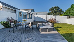 Un patio exterior moderno con una mesa de comedor y asientos, rodeado por una cerca blanca y follaje.