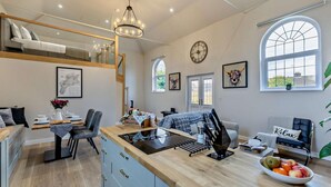Une cuisine moderne avec un îlot central, un coin repas doté d’une table en bois, et une chambre à l’étage, aménagée en mezzanine.