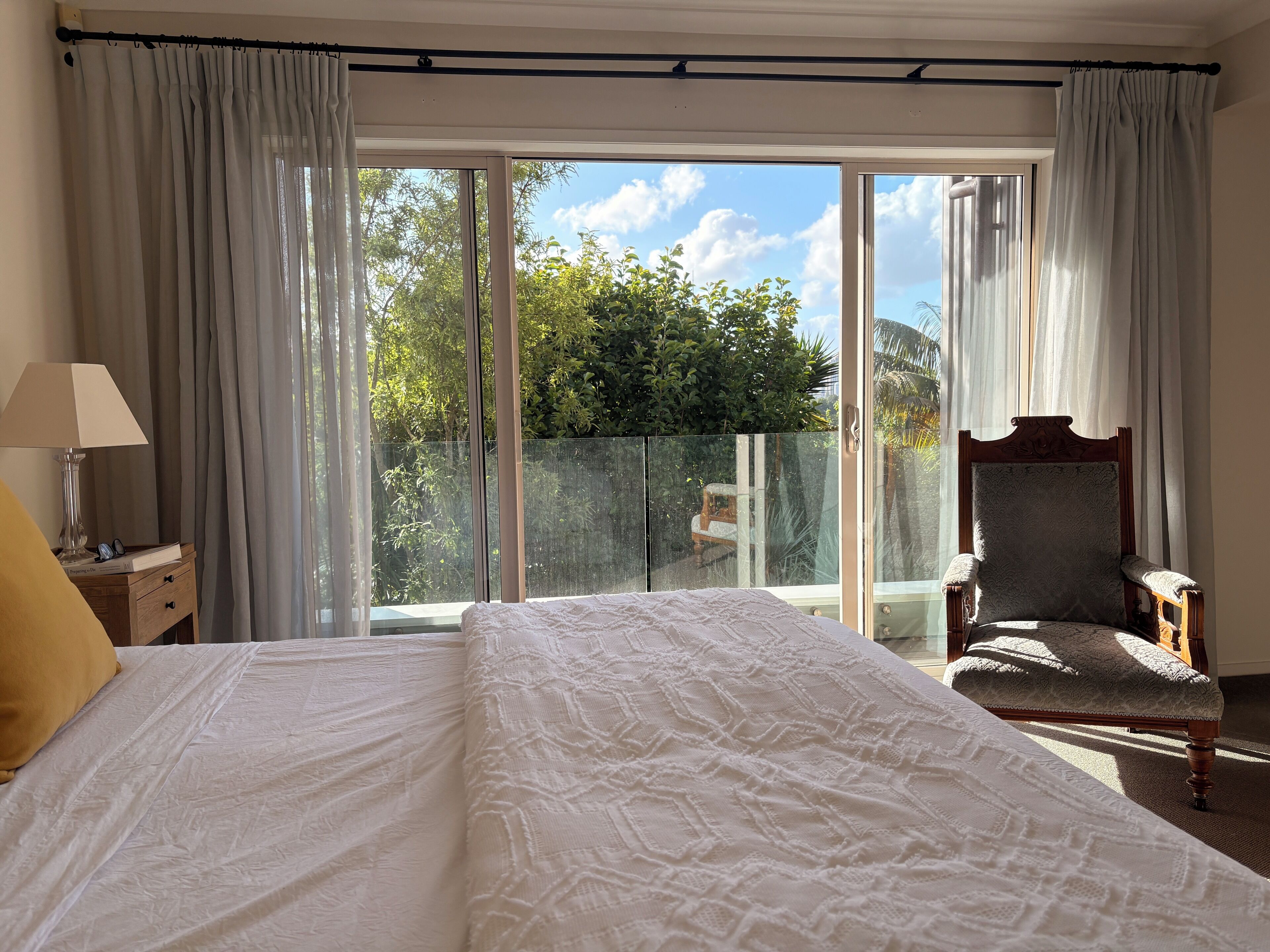 A bedroom with a large bed, a chair, a nightstand, and a view of a balcony with a glass door.