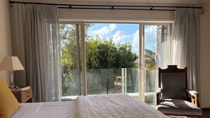 Quarto com cama grande, uma cadeira, mesa de cabeceira e vista para uma varanda com porta de vidro.