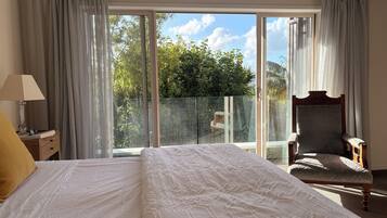 A bedroom with a large bed, a chair, a nightstand, and a view of a balcony with a glass door.