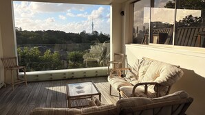 Uma varanda com móveis de vime, uma mesa de vidro e vista para a paisagem urbana e uma torre.