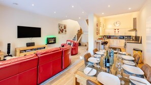 A modern living room with a red sofa, a flat-screen TV, and a dining area with a glass table set for six. - 3 Bed in Torquay (oc-t29804) (Torquay)