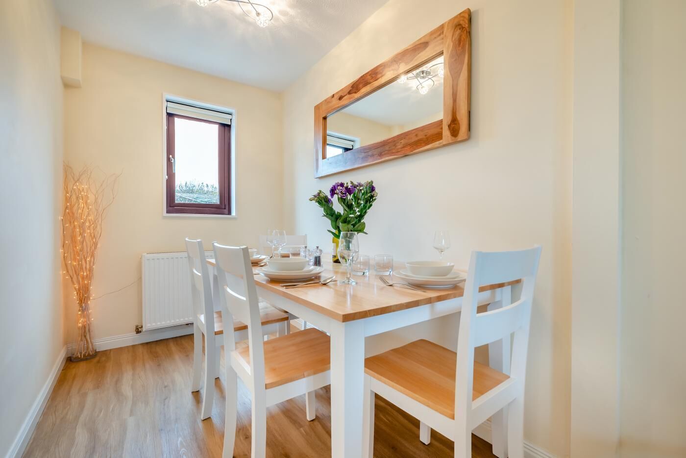 A dining area with a wooden table, white chairs, a vase with flowers, and a mirror on the wall.