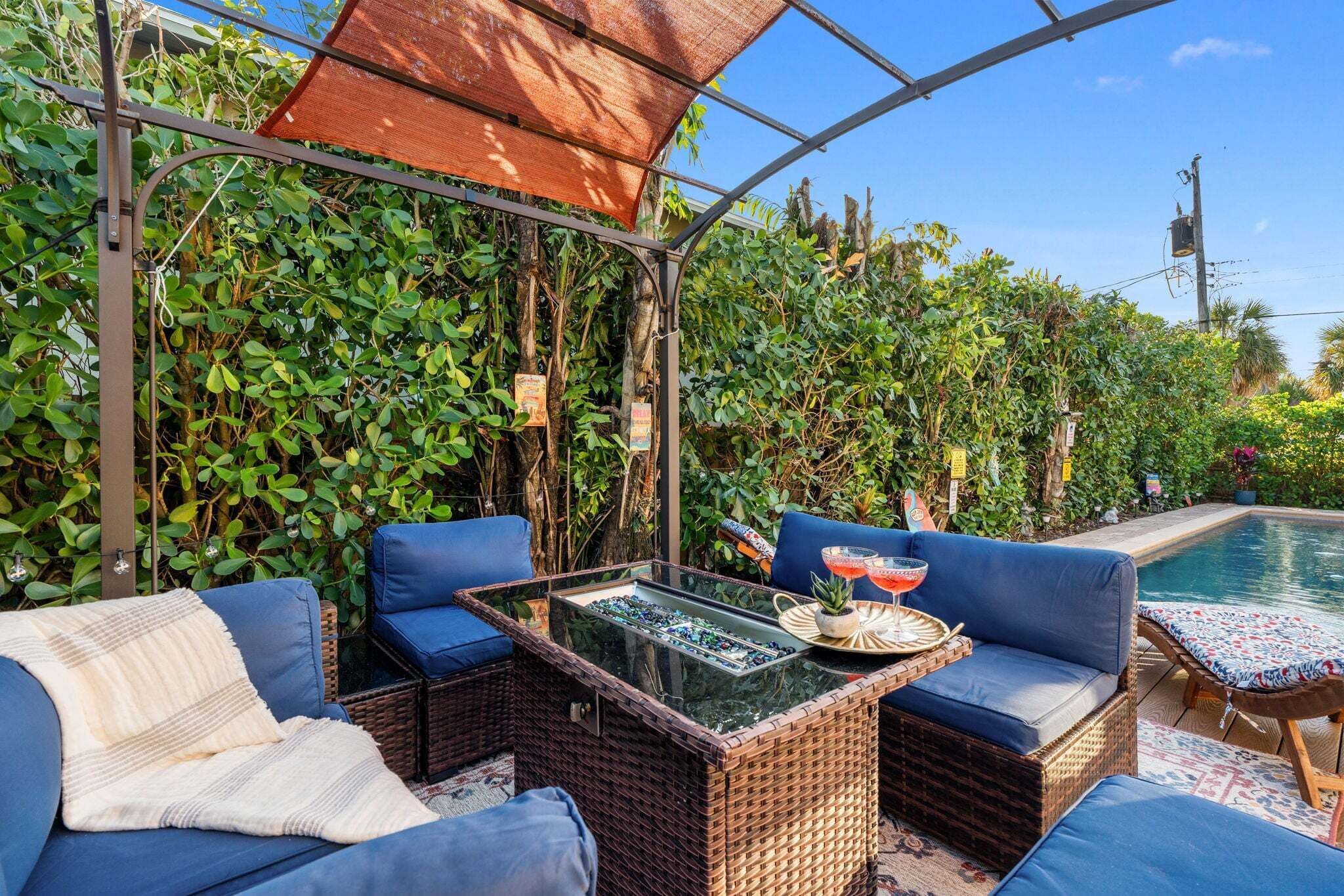 Un espacio de estar al aire libre techado con mobiliario de mimbre, una mesa de vidrio y una piscina al fondo.