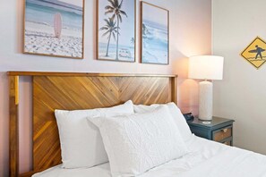 Quarto de hotel com cabeceira de madeira, roupa de cama branca, luminária de cabeceira e decoração de parede com tema de praia.