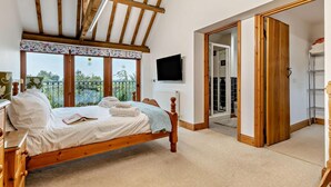 A bedroom with a wooden bed, a TV mounted on the wall, and a view of the outdoors through large windows. - 4 Bed in Litcham (oc-h30106) (Litcham)