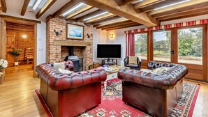 A cozy living room with a brick fireplace, leather sofas, a flat-screen TV, and a large window with a view of the garden. - 4 Bed in Litcham (oc-h30106) (Litcham)