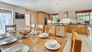 A modern kitchen with wooden cabinets, a central island, and a dining area set for six with a table, chairs, and a fruit bowl. - 4 Bed in Ellastone (oc-11744sf) (Ellastone)