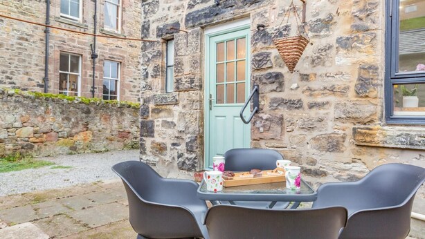 A stone building with a green door, a hanging basket, and a table with chairs and a tray of muffins. - 2 Bed in Spittal (oc-b30119) (Spittal)
