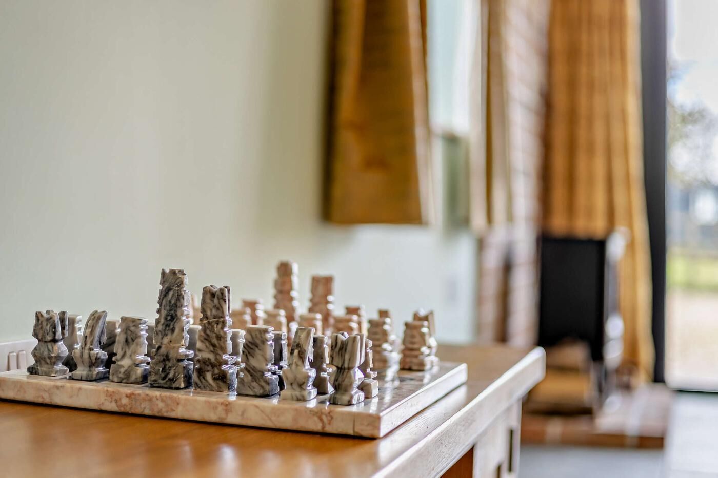 A marble chessboard with pieces on a wooden table in a room with a large window and curtains.