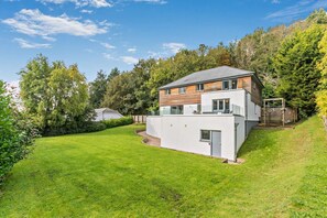 Uma casa moderna com dois andares, um amplo jardim frontal, um deck de madeira e uma pequena área de jardim.