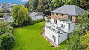 Une maison moderne à deux étages, dotée d’un toit plat, d’un balcon avec des meubles d’extérieur et d’une pelouse bien entretenue.