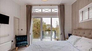 Une chambre à coucher comprenant un lit, une chaise, un téléviseur et donnant sur un lac grâce à une porte vitrée.