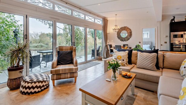 Un vaste espace de vie comprenant un canapé, un fauteuil et un pouf, une table basse en bois et une vue sur un jardin grâce à de grandes fenêtres.