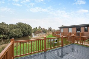 A wooden deck with a view of a river and greenery, a picnic table, and a building with windows. - 2 Bed in Welney (oc-c30124) (Welney)