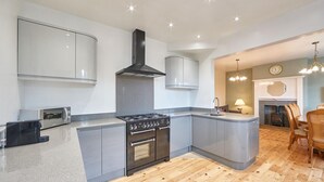 Een moderne keuken met een gasfornuis, oven en inbouwmeubels. Er is een zithoek met houten vloeren en een open haard.