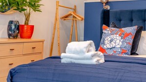 Un lit bien fait, recouvert d’une couvre-lit bleue, une commode en bois avec des tiroirs, un vase rouge, une plante en pot, un cintre en bois et une tête de lit bleue ornée d’un coussin à motifs floraux.
