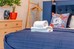 A neatly made bed with a blue bedspread, a wooden dresser with drawers, a red vase, a potted plant, a wooden hanger, and a blue headboard with a floral pillow. - 4 Bed in Carbis Bay (oc-r30048) (Carbis Bay)