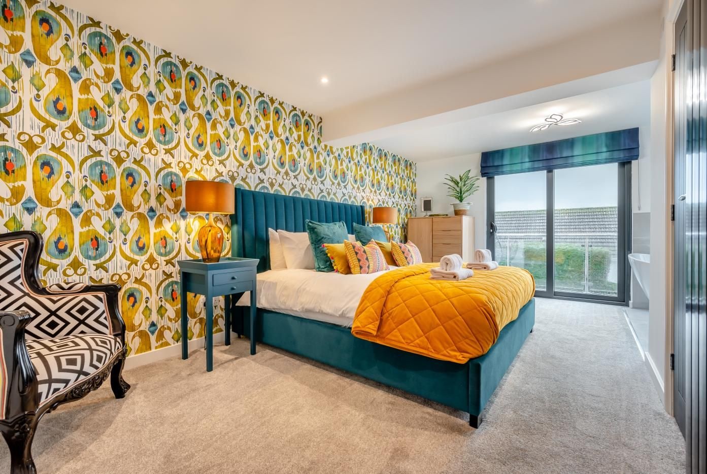 A bedroom with a large bed, a nightstand, a chair, a dresser, and a view of the outside.
