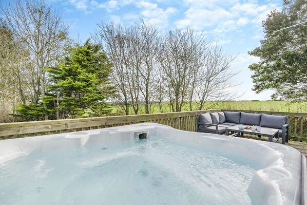A hot tub with a wooden deck, a table with cushions, and a view of trees and open fields.