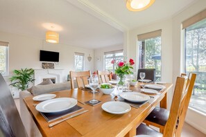 A dining area with a wooden table set for a meal, chairs, a television on the wall, and a window with a view of trees.