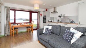 A cozy living room with a grey sofa, wooden dining table, and a view of the sea through large windows. - 1 Bed in Reef (oc-r30134) (Uig)