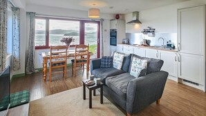 A cozy living room with a sofa, a dining table with chairs, and a view of the sea through large windows. - 1 Bed in Reef (oc-r30134) (Uig)