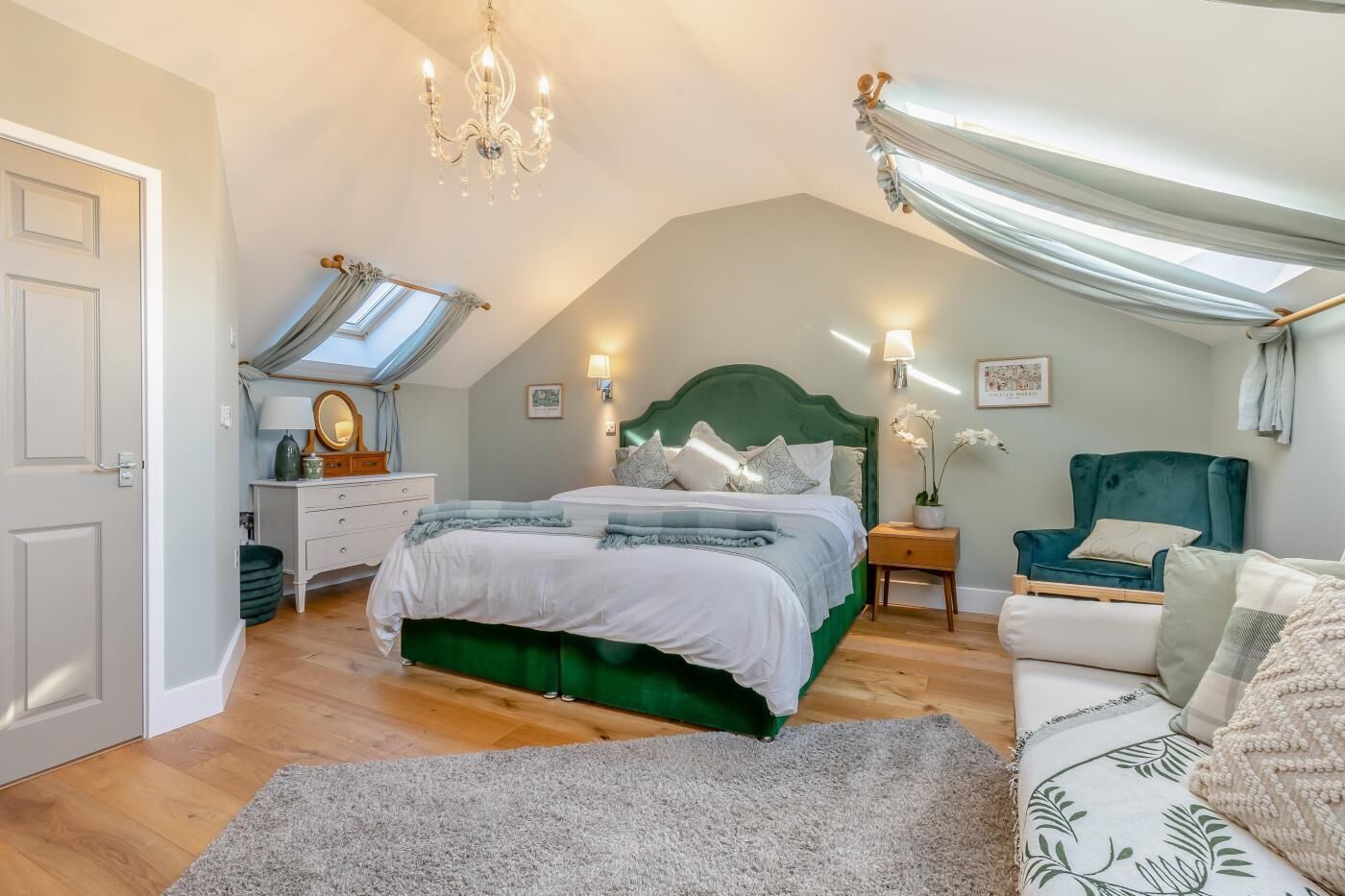 Ein Schlafzimmer mit einem großen Bett, einem grünen Kopfteil, einem Nachttisch, einer Kommode, einem Sessel und einer Deckenleuchte.