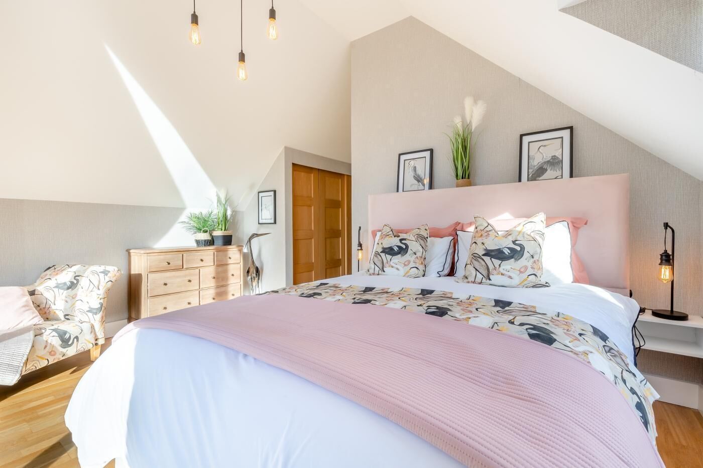 A bedroom with a large bed, a wooden dresser, a desk with a lamp, and a view of the outdoors.