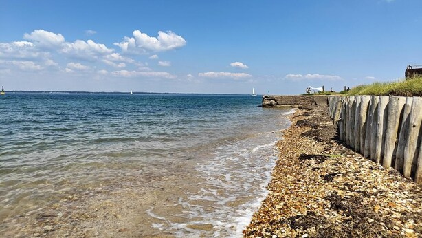 Una playa costera con aguas cristalinas, un litoral rocoso y un faro en la distancia.