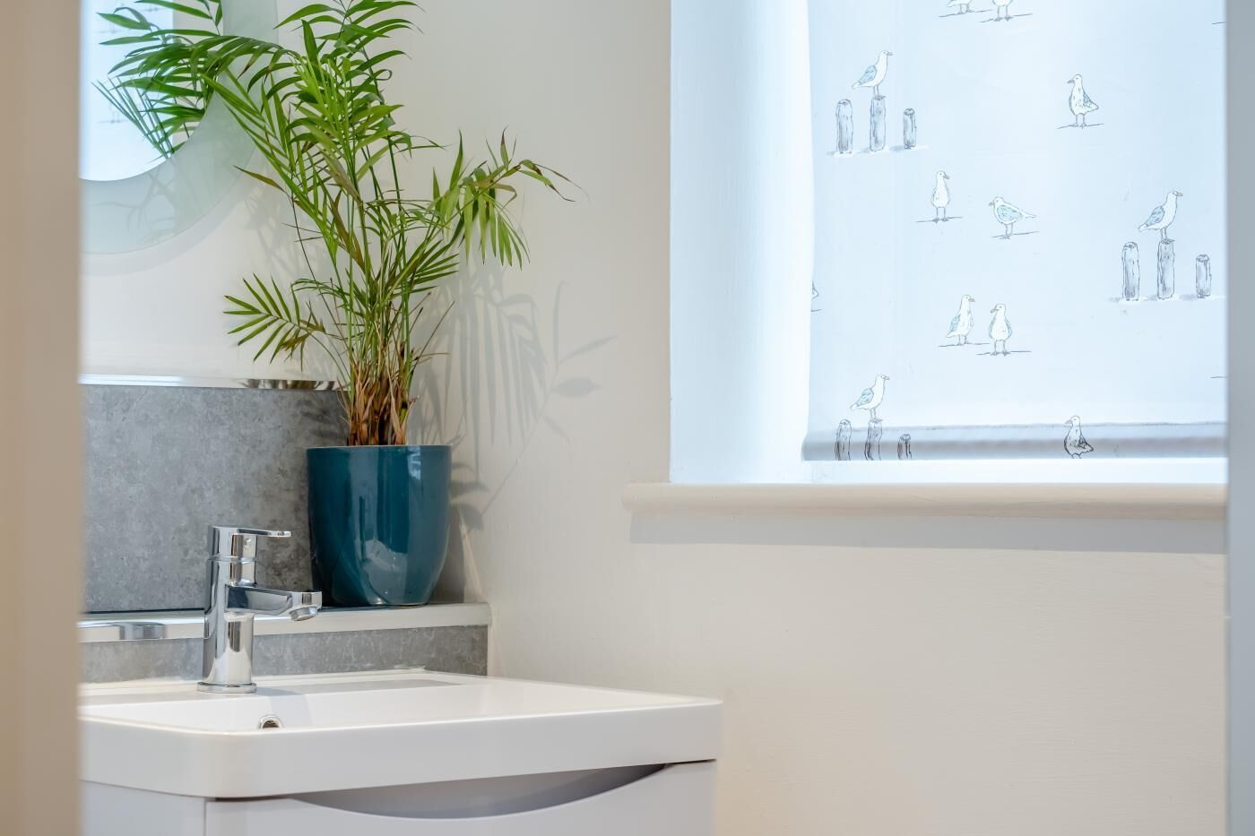 A bathroom with a white sink, a blue vase with green plants, and a window with a bird pattern.