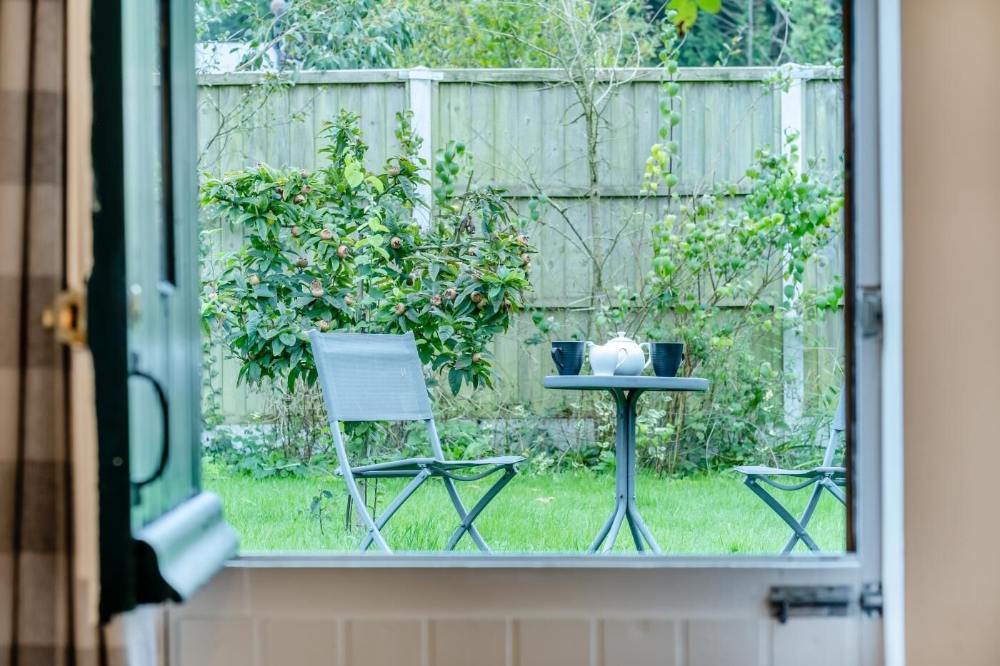 A view from a window to a patio with a table, two chairs, and a small garden.