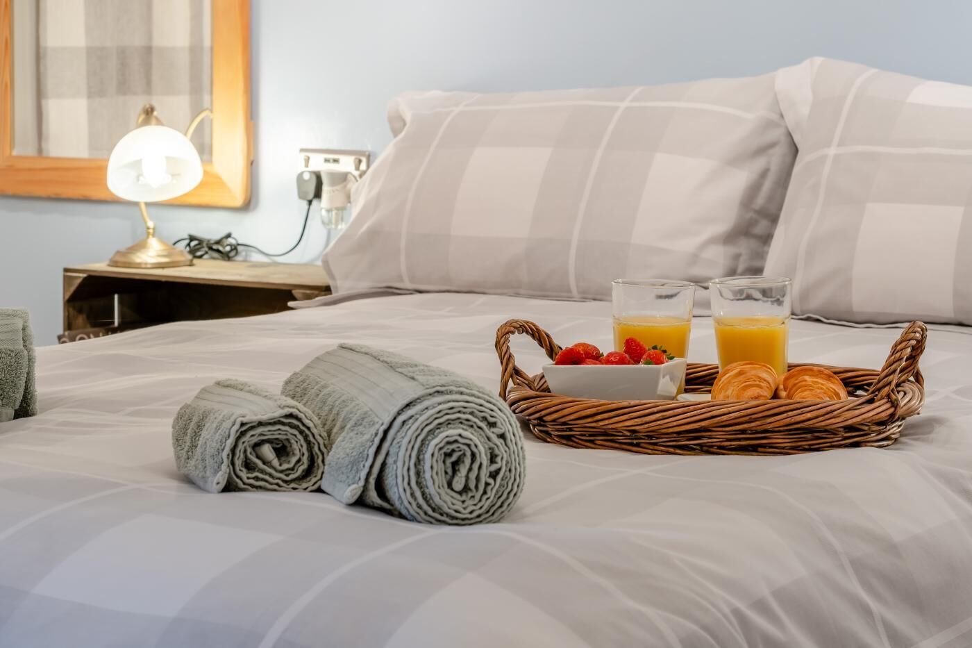 A neatly made bed with rolled towels, a basket of breakfast including croissants, strawberries, and orange juice, and a bedside lamp.