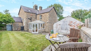 A stone house with a conservatory, a wooden table with chairs, and a grassy yard. - 2 Bed in Aldbrough St. John (oc-l30130) (Aldbrough Saint John)